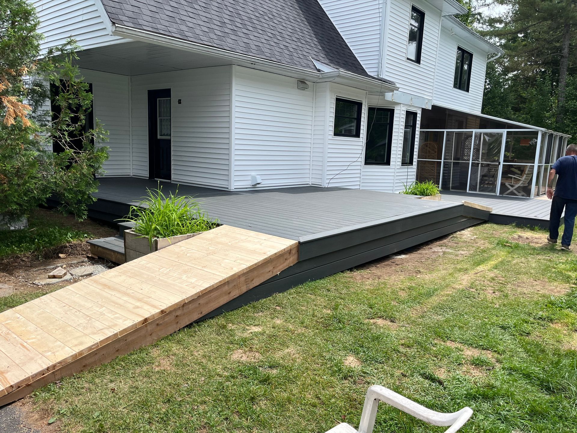 Maison blanche avec terrasse grise et rampe en bois. Une personne se tient près de la véranda. Pelouse verte.