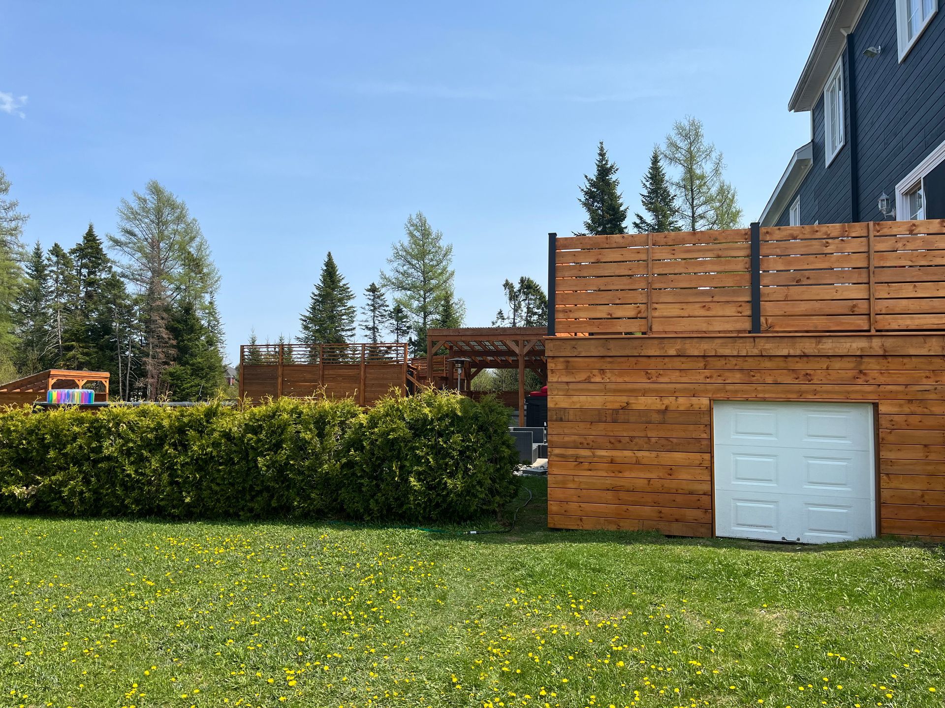 Clôture en bois et porte de garage dans une cour arrière avec pelouse verte, arbres et ciel bleu.