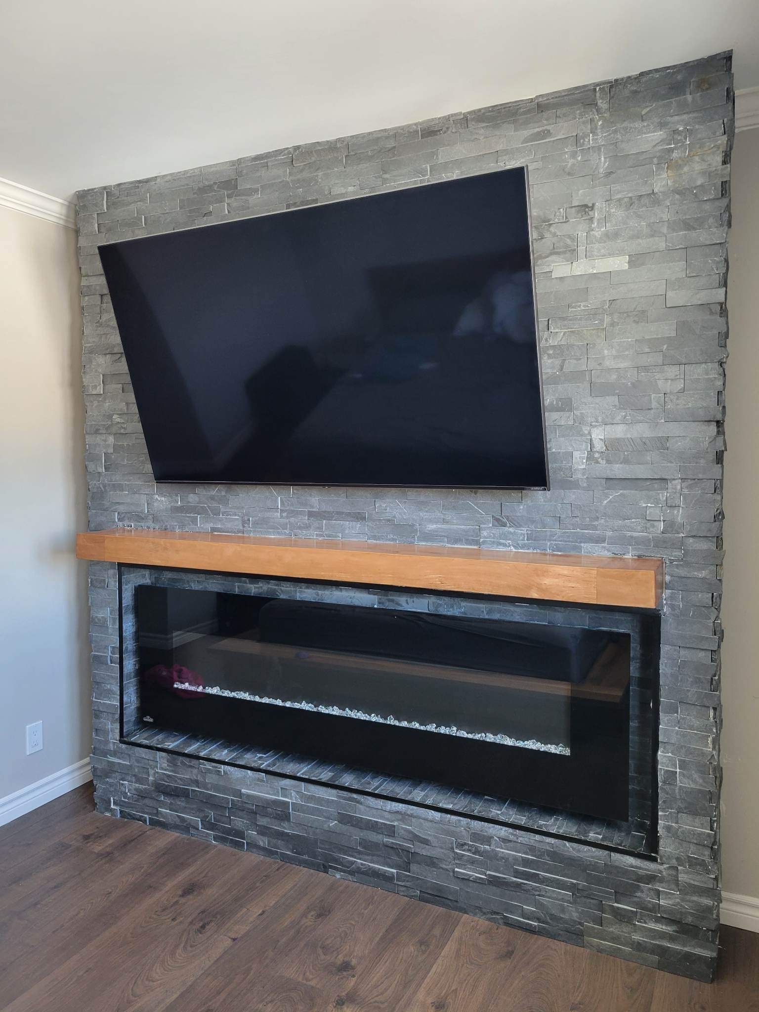 Cheminée avec un téléviseur noir fixé au-dessus, sur un mur carrelé d'ardoises et surmonté d'un manteau en bois.