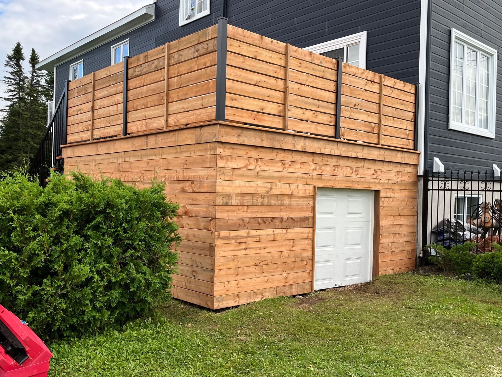 Terrasse en bois au-dessus d'un garage aux murs bardés de bois horizontaux. On aperçoit une porte de garage blanche.
