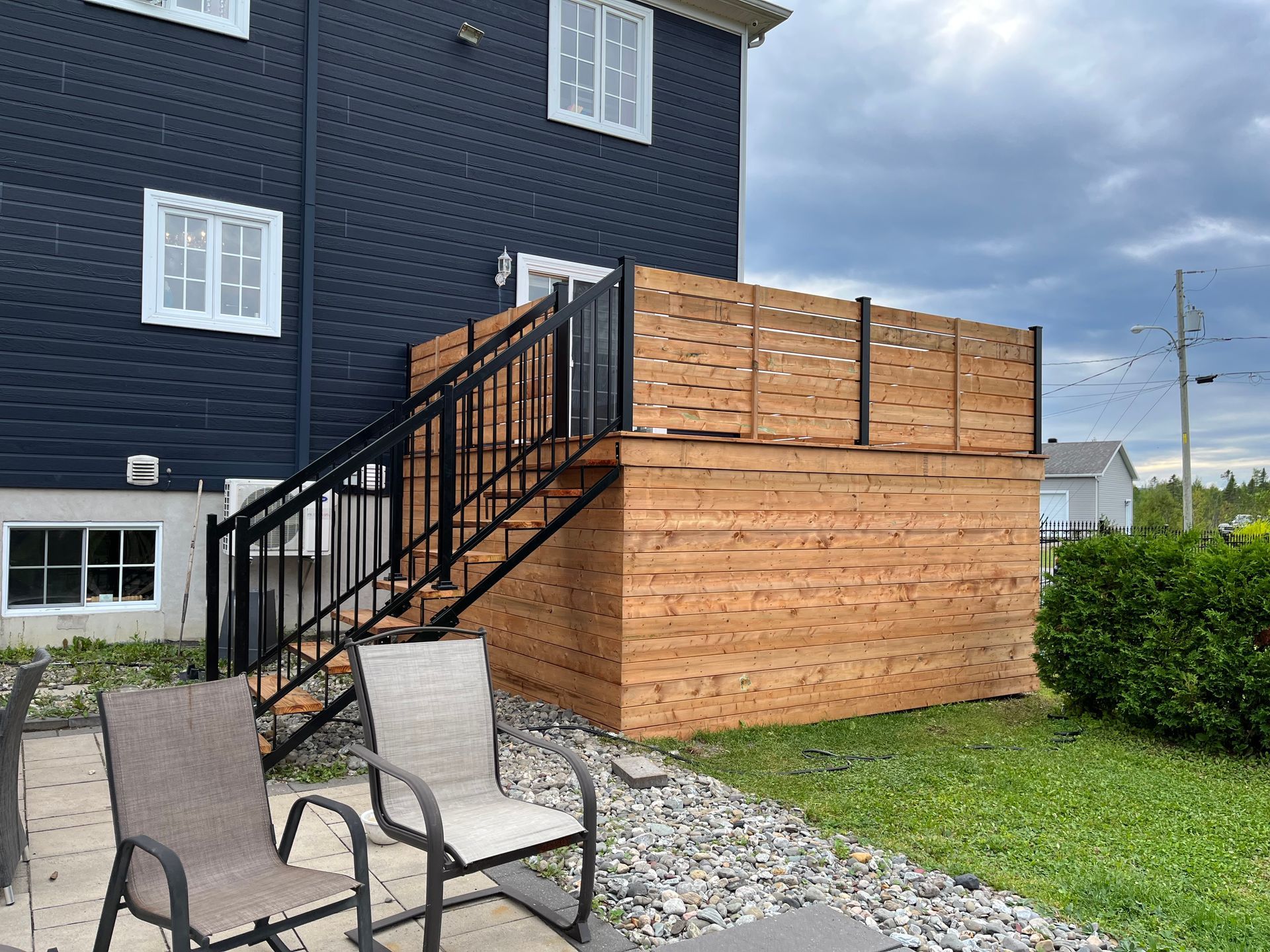 Terrasse en bois avec rambarde noire et clôture préservant l'intimité, attenante à une maison bleu foncé. Deux chaises sont disposées sur le patio.