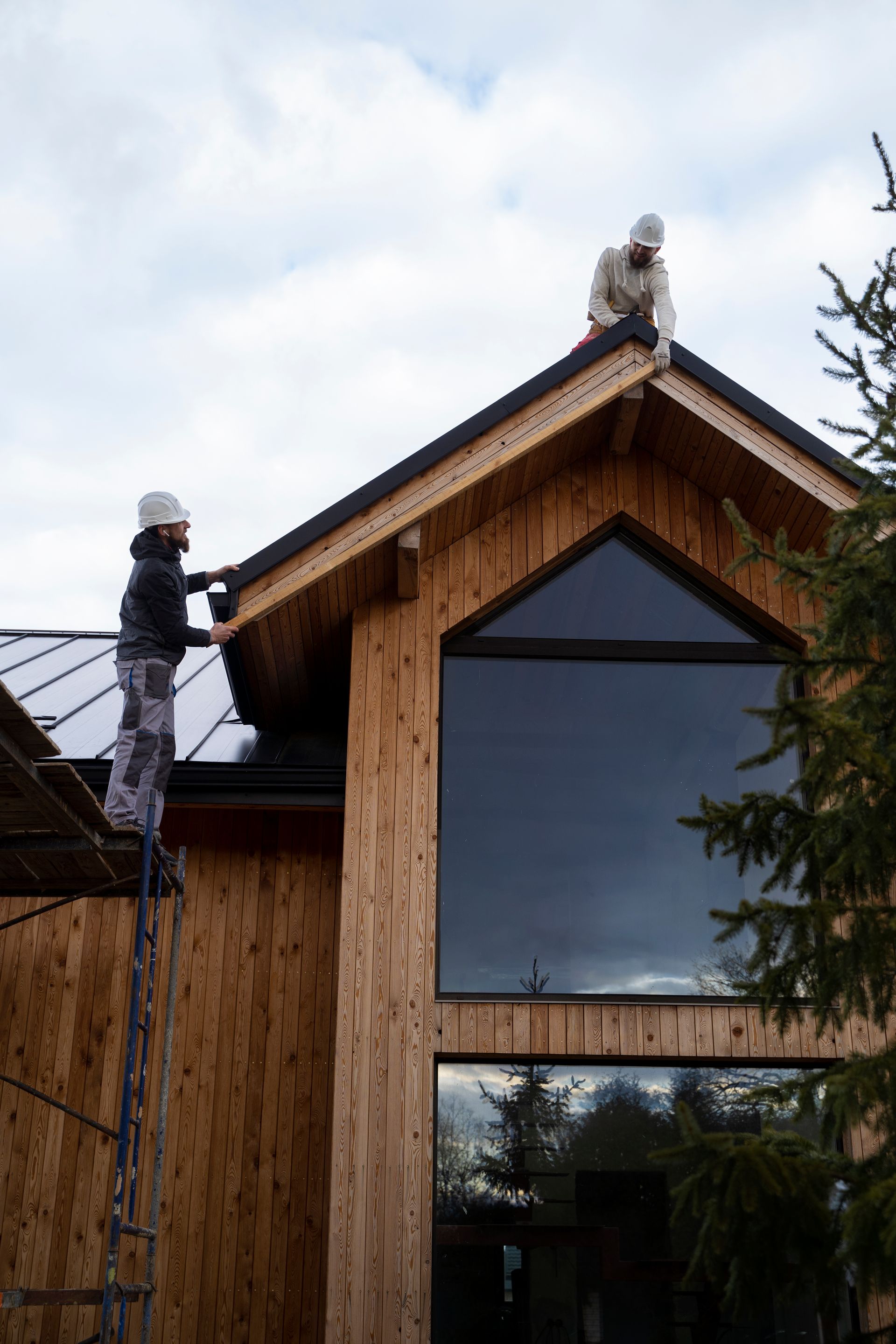 Des ouvriers du bâtiment installent des matériaux sur le toit d'une maison en bois, avec de grandes fenêtres, sous un ciel nuageux.