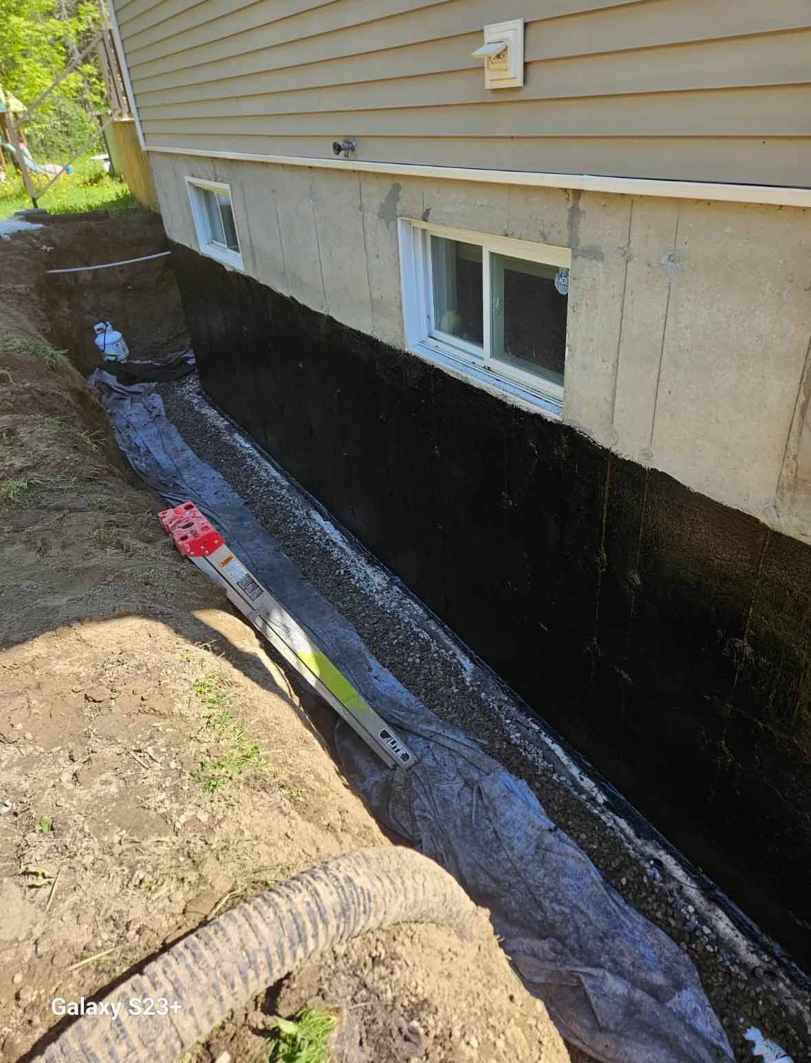Étanchéité des fondations : Membrane noire appliquée sur un mur en béton dans une tranchée, le long d'une fenêtre blanche et d'un bardage beige.