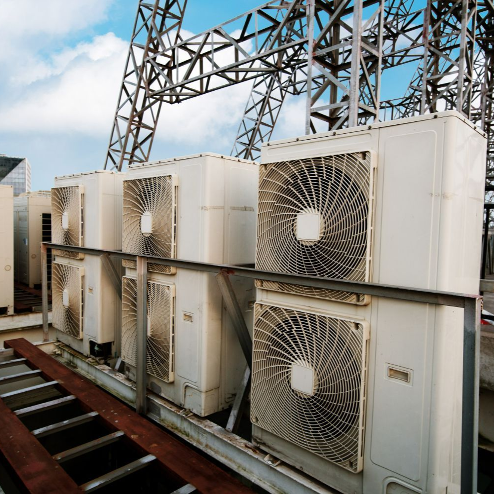 Row of white air conditioning units on a rooftop with metal framework and a blue sky in the background.