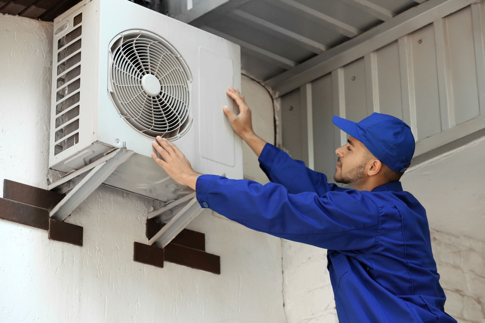 Man in blue uniform installing an air conditioning unit on a white brick wall.