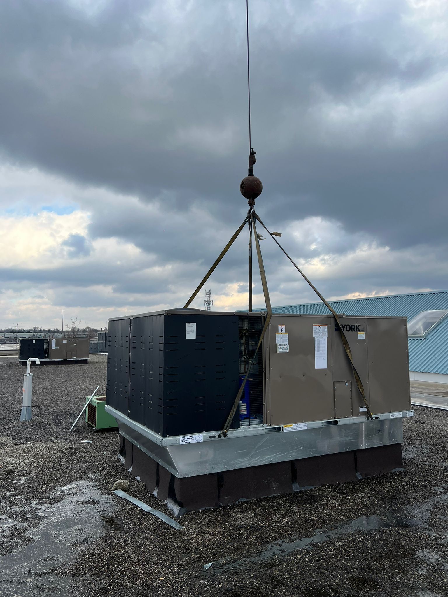 Rooftop crane lifting a large HVAC unit with dark metal exterior, silver base, and tan box on top. Cloudy sky.