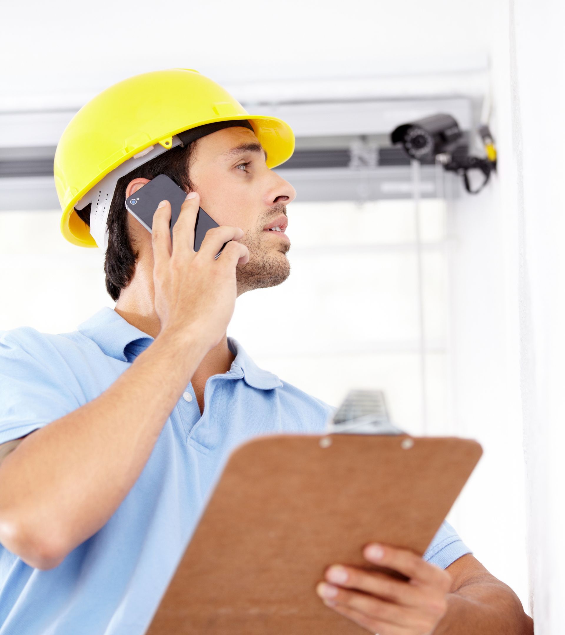 Man in yellow hard hat on phone, inspecting security camera, holding clipboard indoors.
