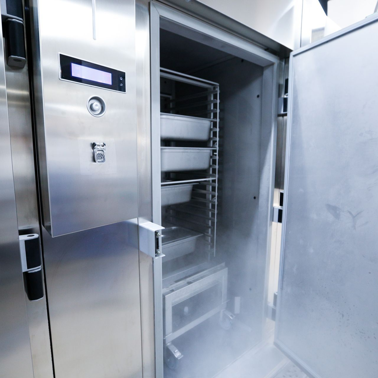 Stainless steel freezer with open door, showing trays and misty interior.