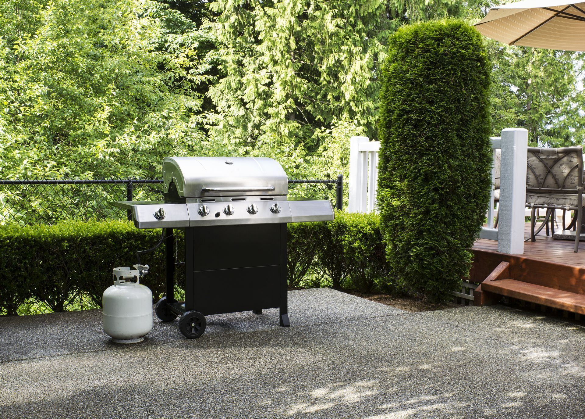 Grill with propane tank on a gravel patio near green shrubs, next to a deck with outdoor seating.