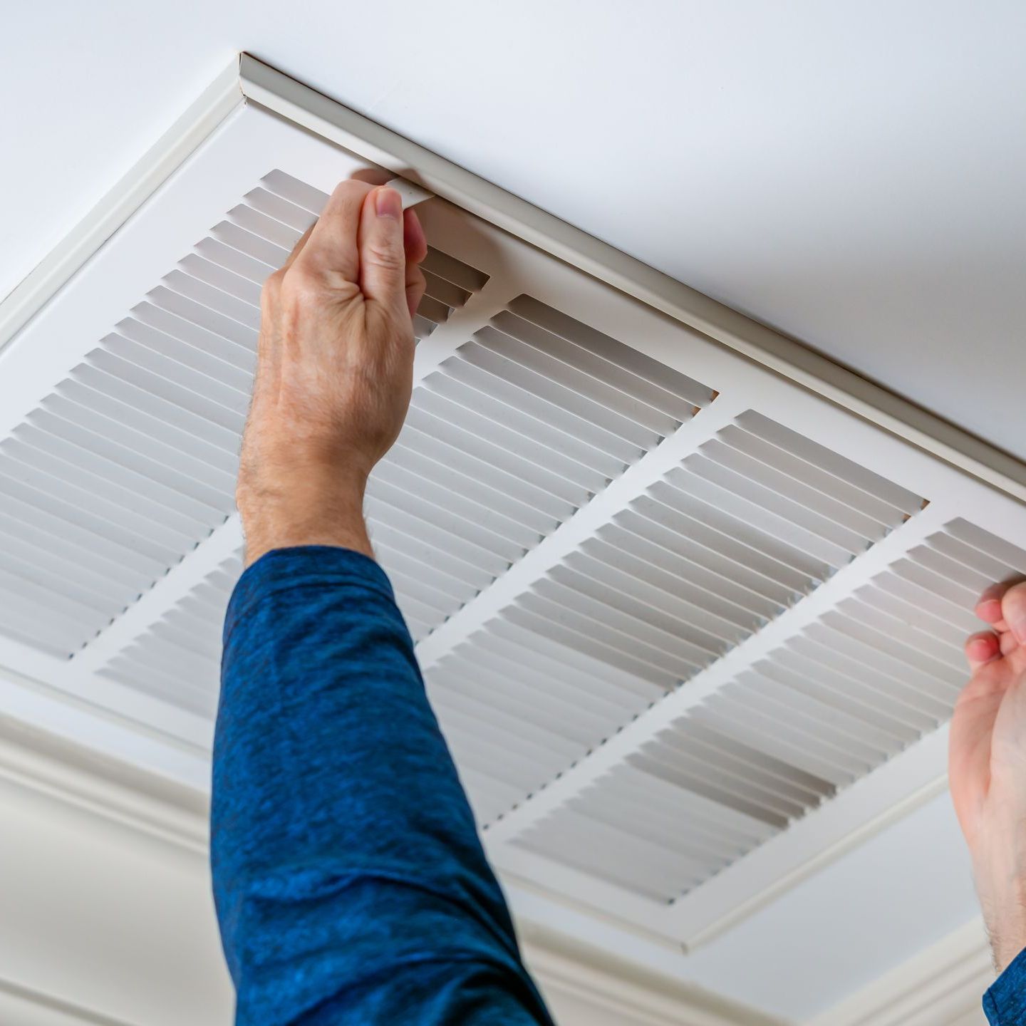 Person removing a ceiling vent cover with hands, white vent, blue shirt.