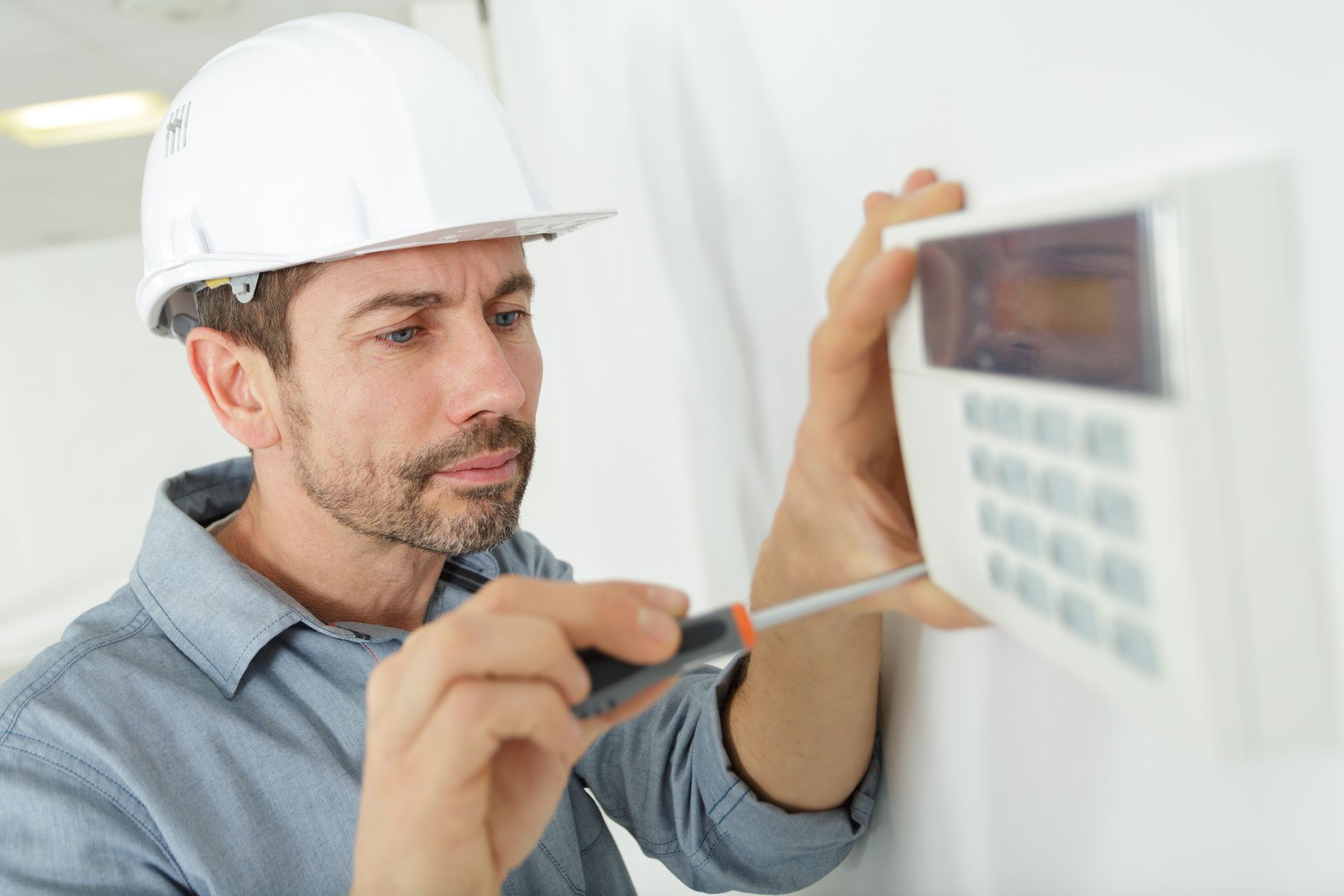 A man in a white hard hat uses a screwdriver to install a white security keypad on a white wall.