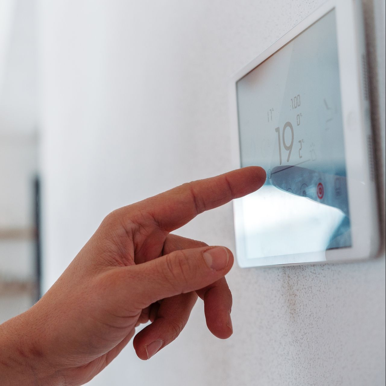 Person's hand touching a digital thermostat on a white wall, adjusting the temperature.