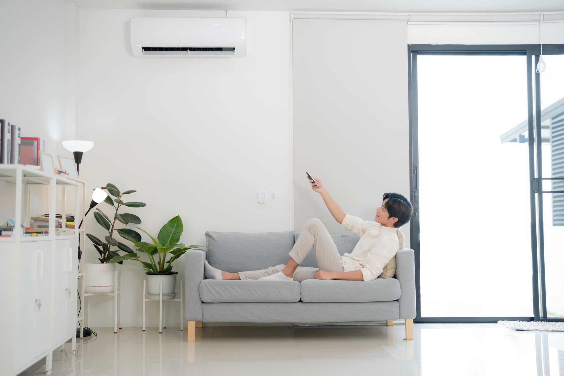 Man on sofa uses remote to adjust wall-mounted air conditioner in a bright, modern living room.
