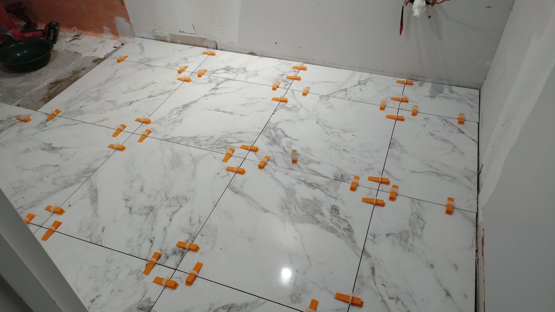 Pose de grands carreaux blancs à motif marbre au sol de la salle de bains ; clips de nivellement orange visibles.