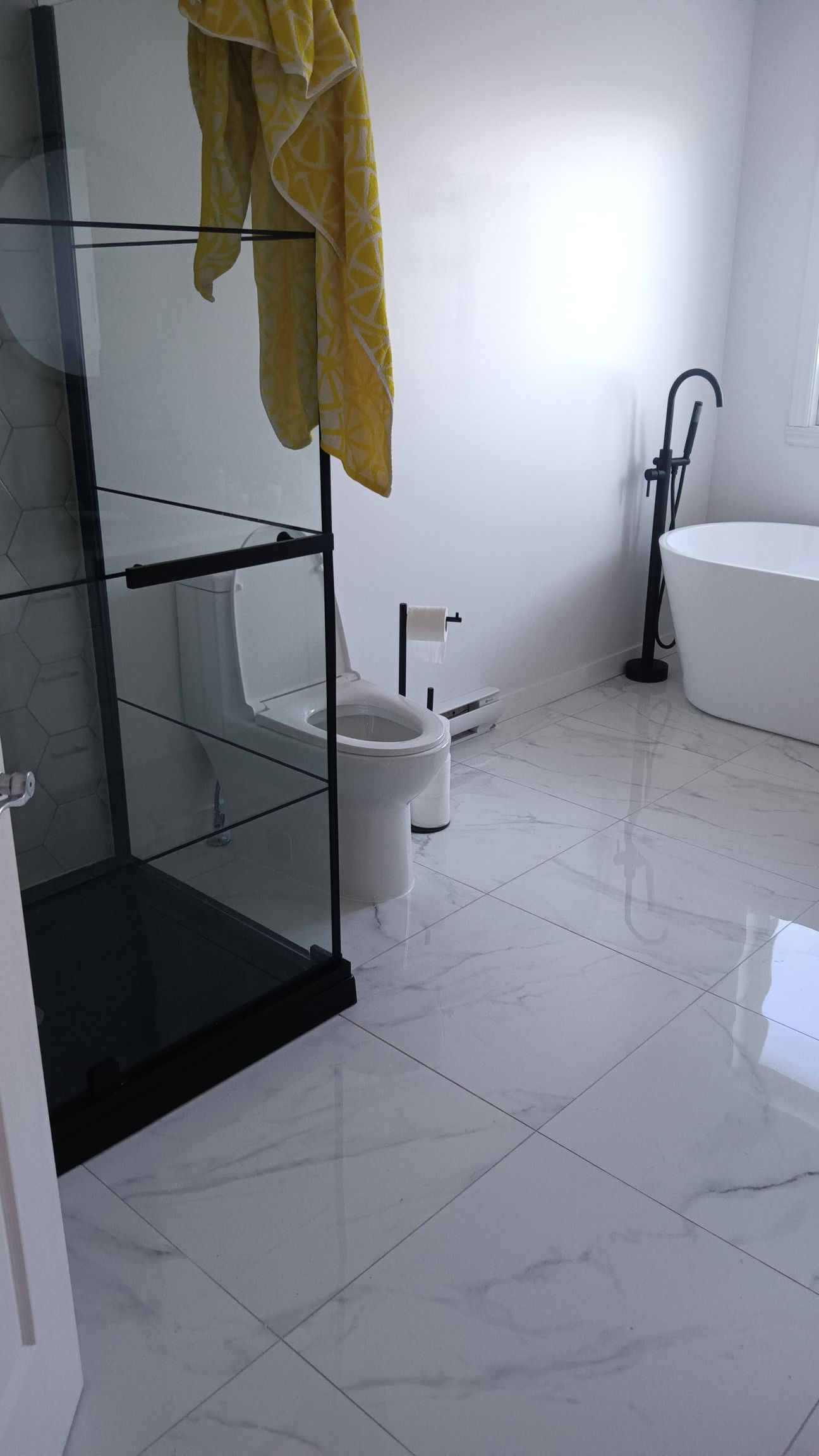 Salle de bains moderne blanche avec sol en marbre, douche noire, toilettes et baignoire îlot. Porte-serviettes jaune.