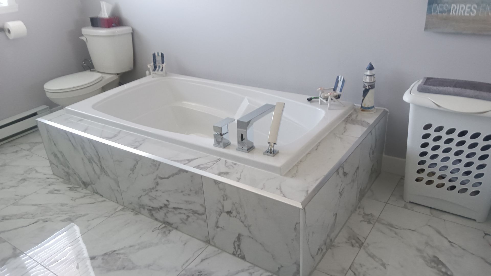 Salle de bains avec baignoire blanche entourée de faux marbre, à côté des toilettes et d'un panier à linge.