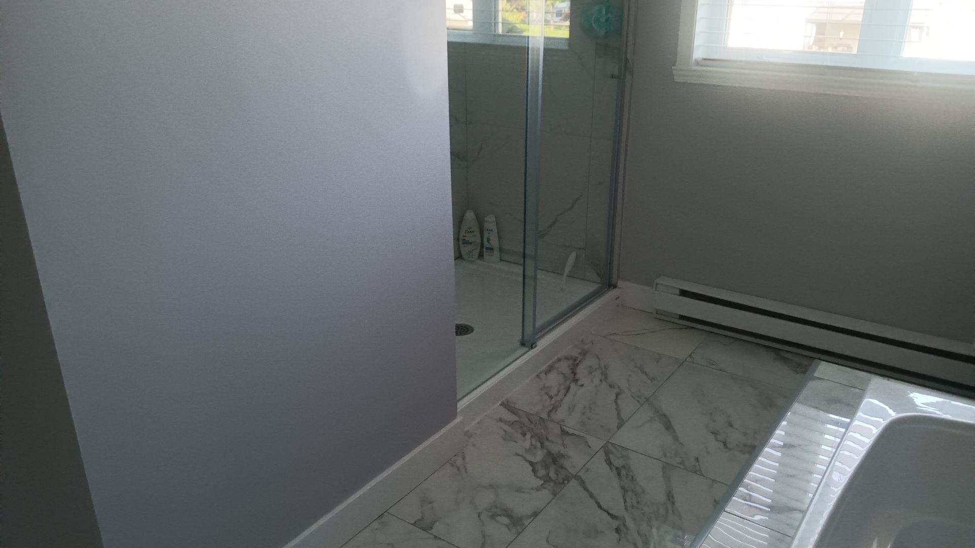Salle de bains avec porte de douche en verre, sol en carrelage de marbre, murs gris et radiateur sous une fenêtre.