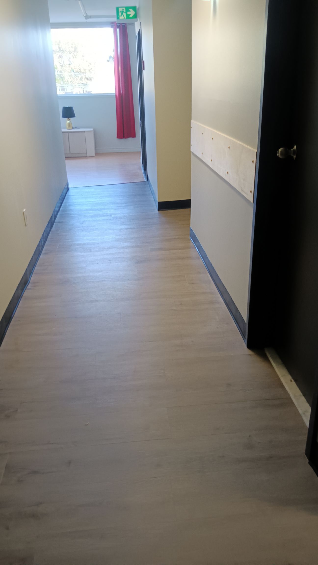 Un couloir étroit au sol clair imitant le bois, menant à une pièce avec une fenêtre et un rideau rouge.