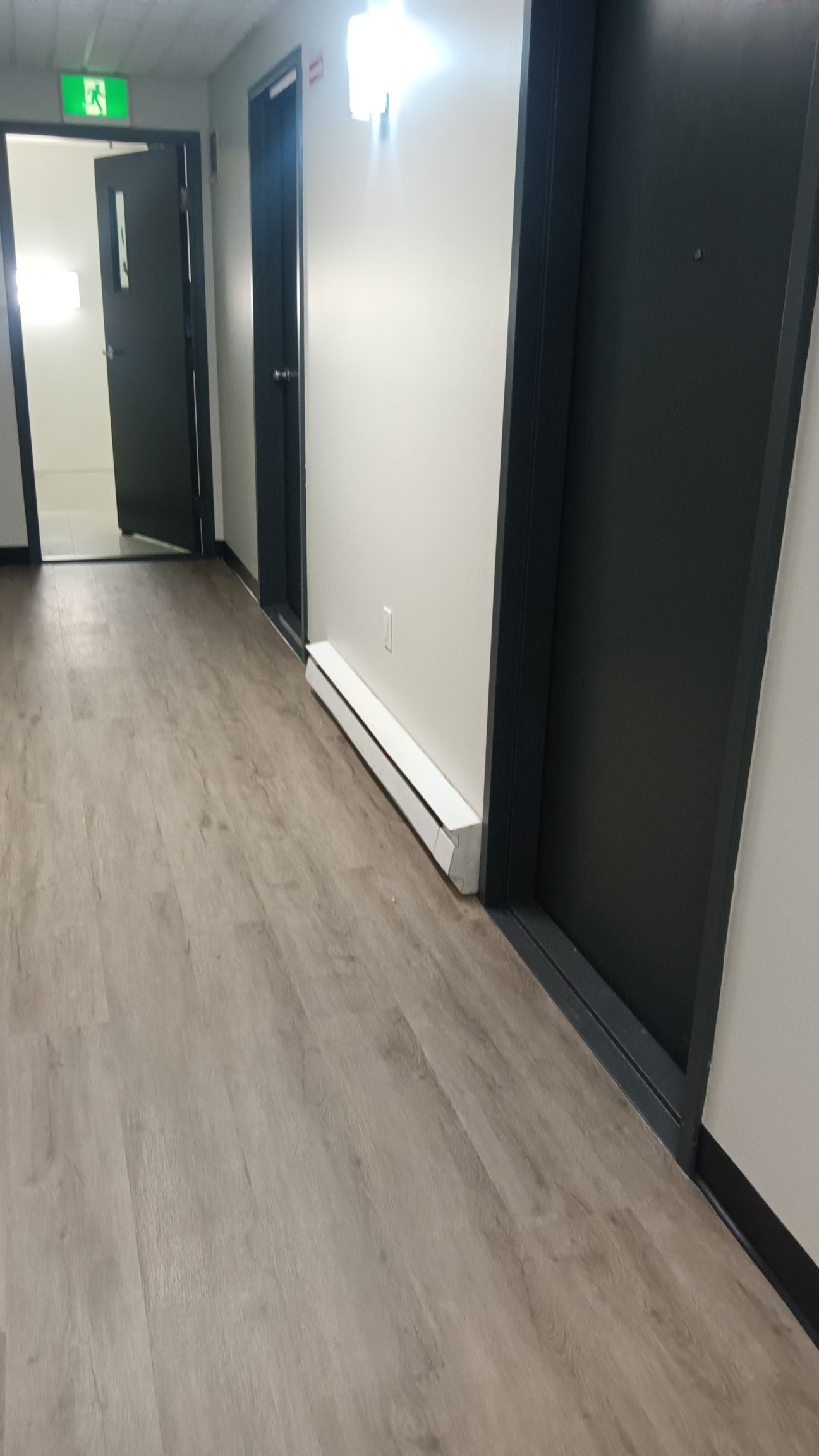 Couloir avec sol imitation bois gris, murs blancs, portes noires et radiateur plinthe. Un panneau de sortie vert est visible.