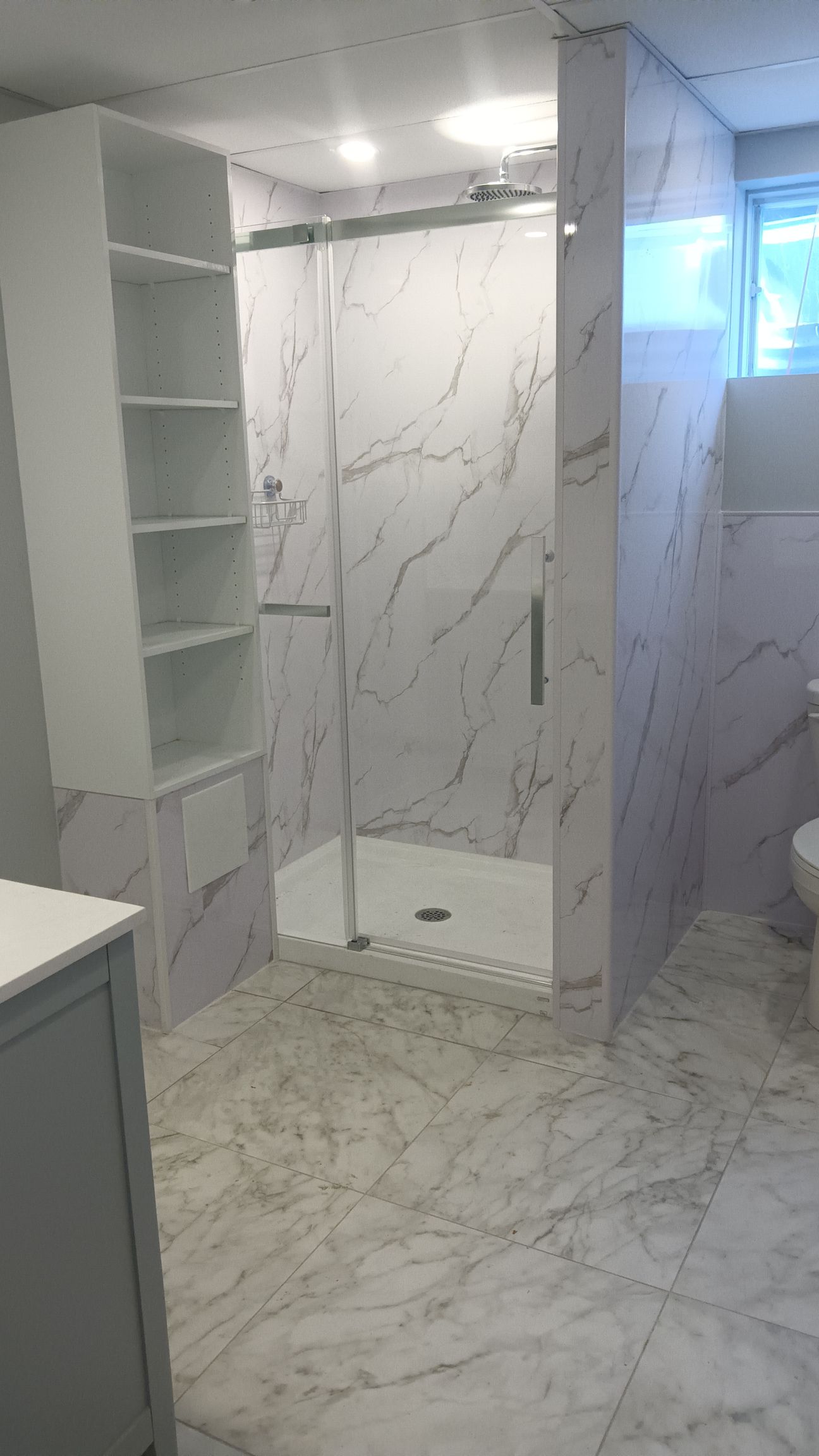 Salle de bains carrelée en marbre avec paroi de douche en verre. Étagères intégrées et toilettes.