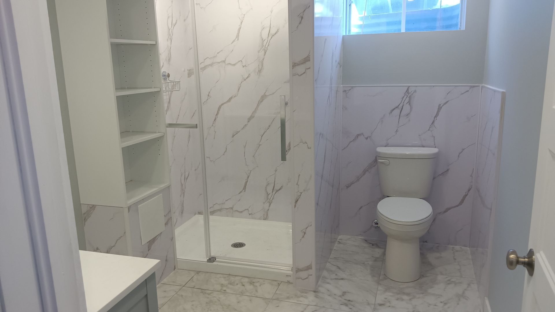 Salle de bains moderne avec carrelage imitation marbre, douche en verre, étagères intégrées et toilettes.