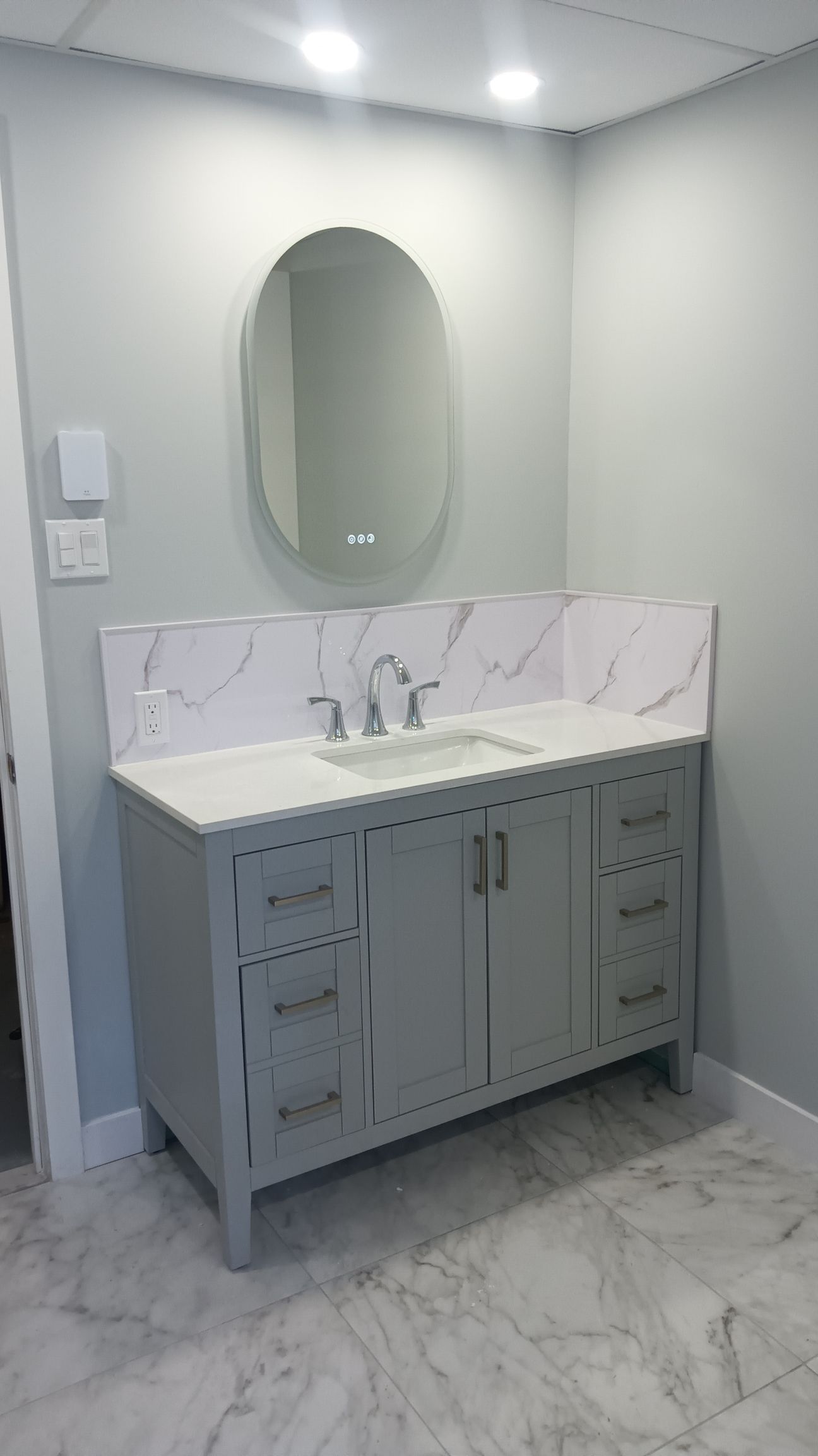 Meuble-lavabo gris avec comptoir en marbre blanc, miroir ovale et murs gris clair.