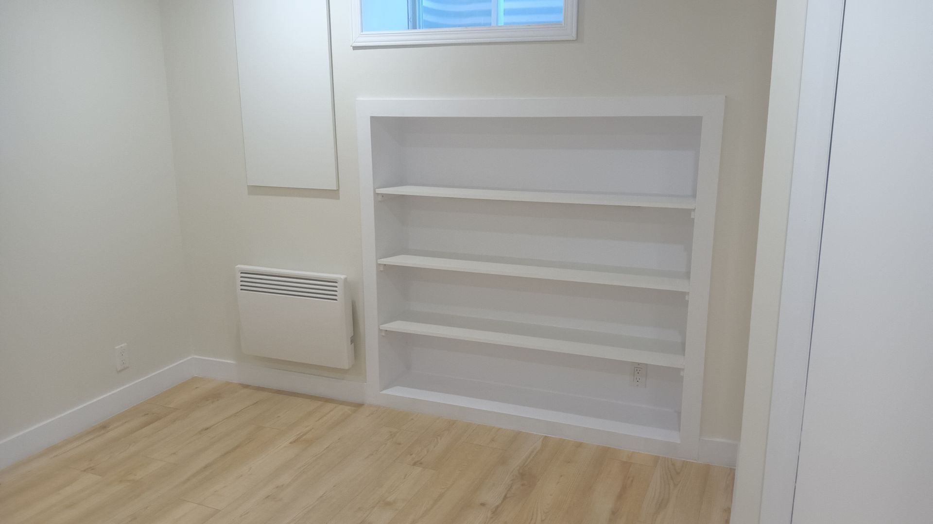 Bibliothèque encastrée blanche vide sur un mur beige dans une pièce au parquet clair.