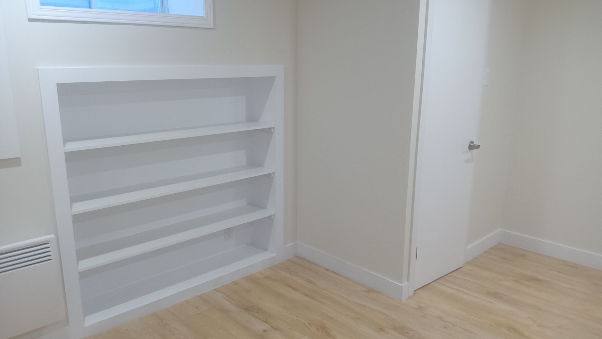 Bibliothèque encastrée blanche à quatre étagères dans une pièce claire avec porte fermée et sol imitation bois.