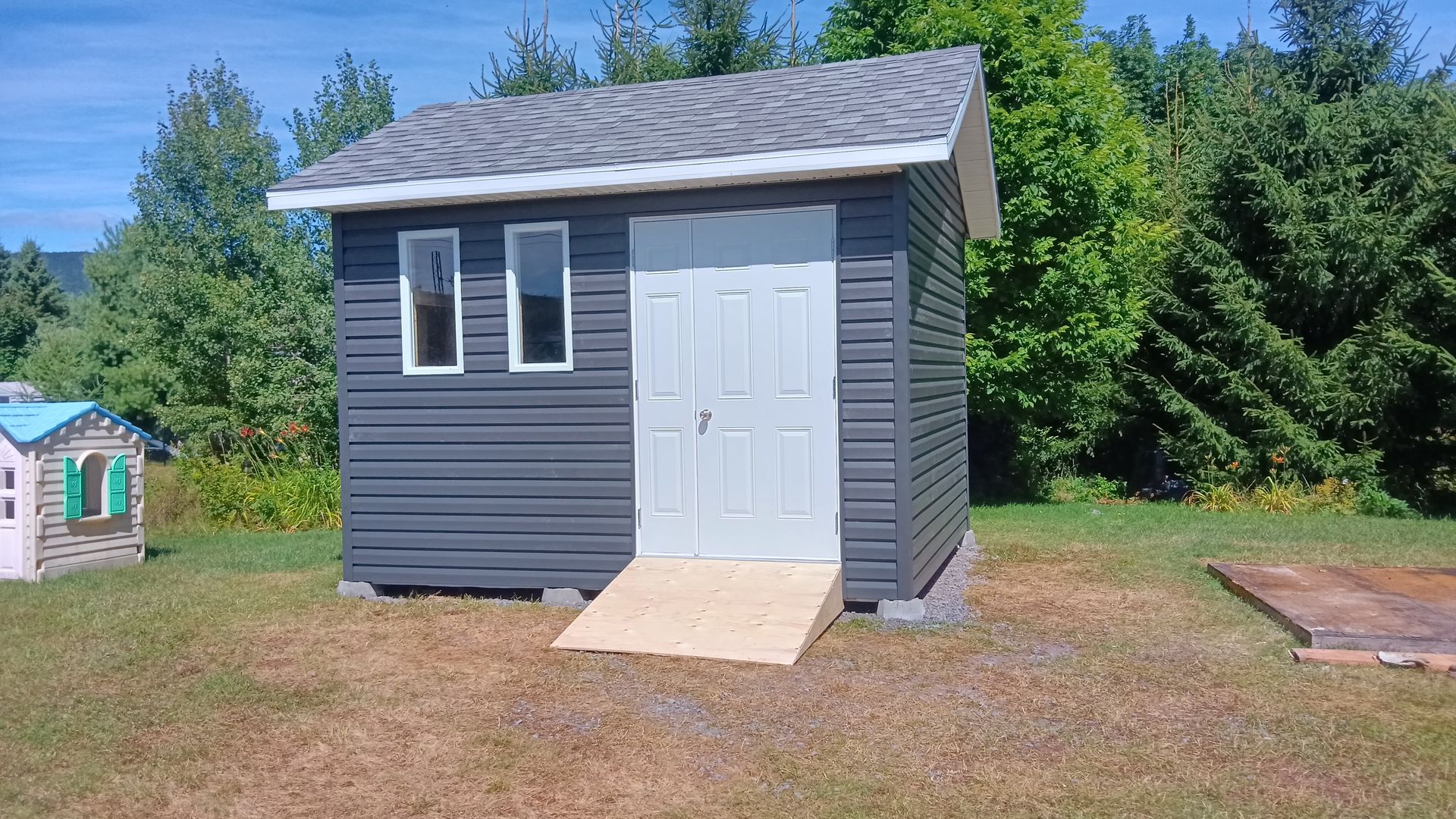Abri gris avec porte blanche, deux fenêtres et rampe d'accès, sur un terrain herbeux.
