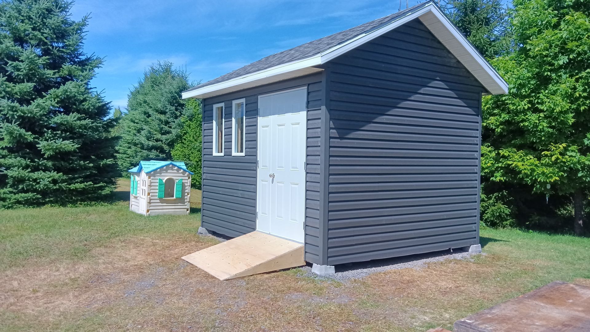 Abri gris foncé avec porte blanche, rampe et petites fenêtres, par une journée ensoleillée.