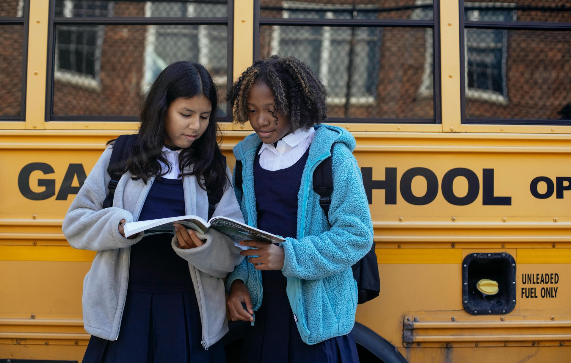 Two girls are reading books in front of a school bus