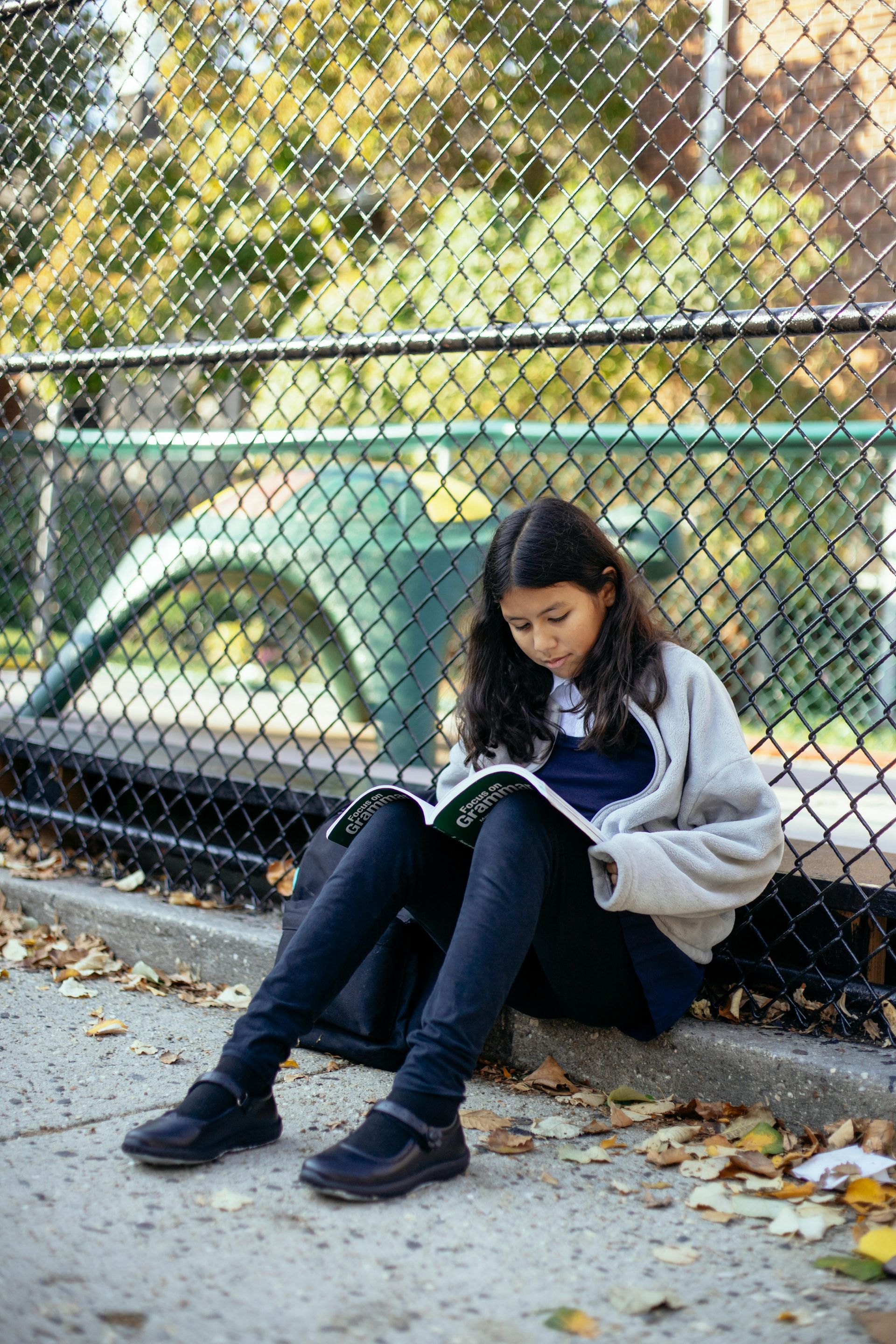 A young girl is sitting on the sidewalk reading a book.