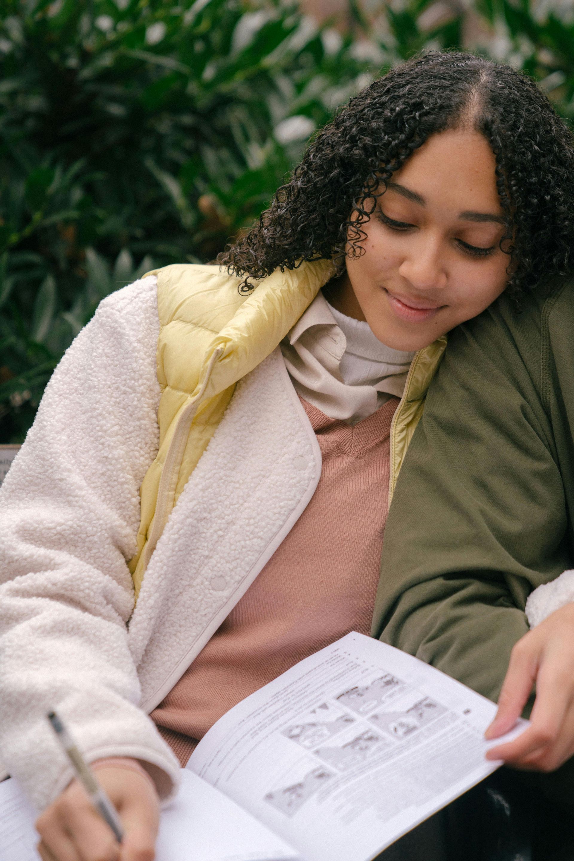 A woman is sitting on a bench reading a book and writing in a notebook.