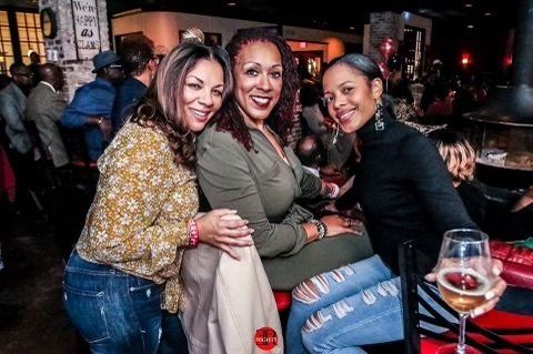Three women are posing for a picture while sitting at a table with a glass of wine.