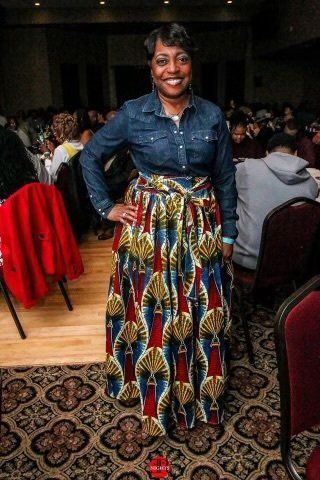 A woman wearing a denim shirt and a long skirt is standing in a room.