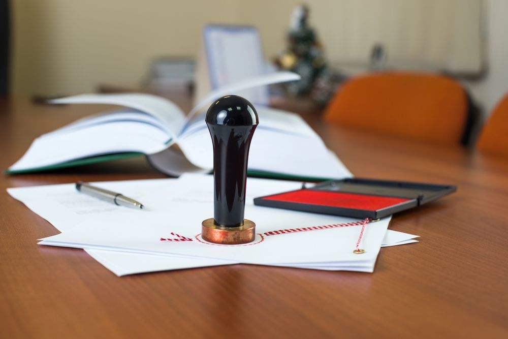 Timbro, tampone e documenti su una scrivania con un libro aperto sullo sfondo.