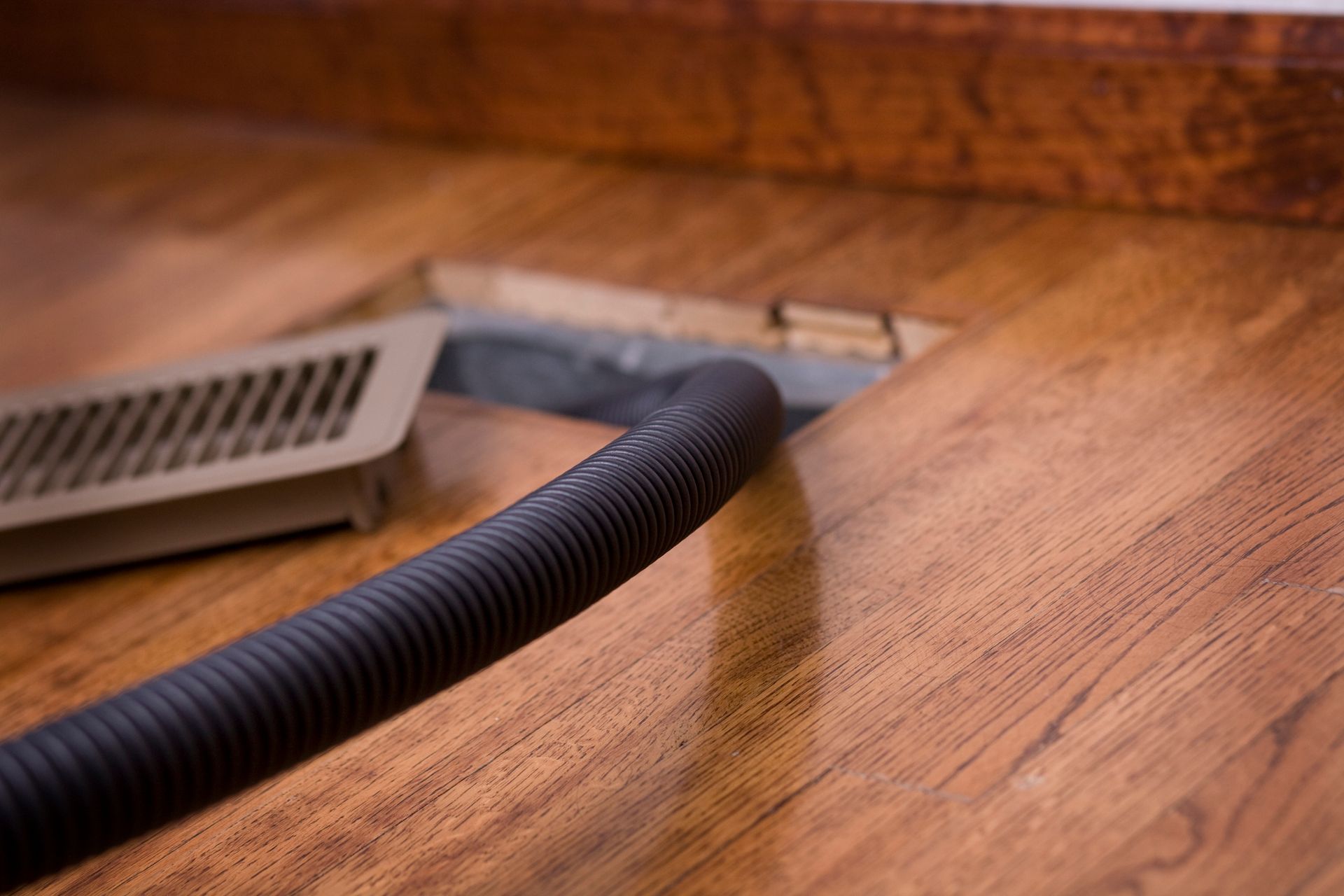 A vacuum hose is being used to clean a vent on a wooden floor.