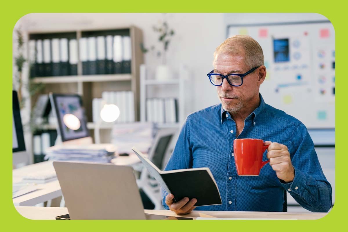 A business man in glasses and a collared shirt drinking coffee and looking at a book in an office