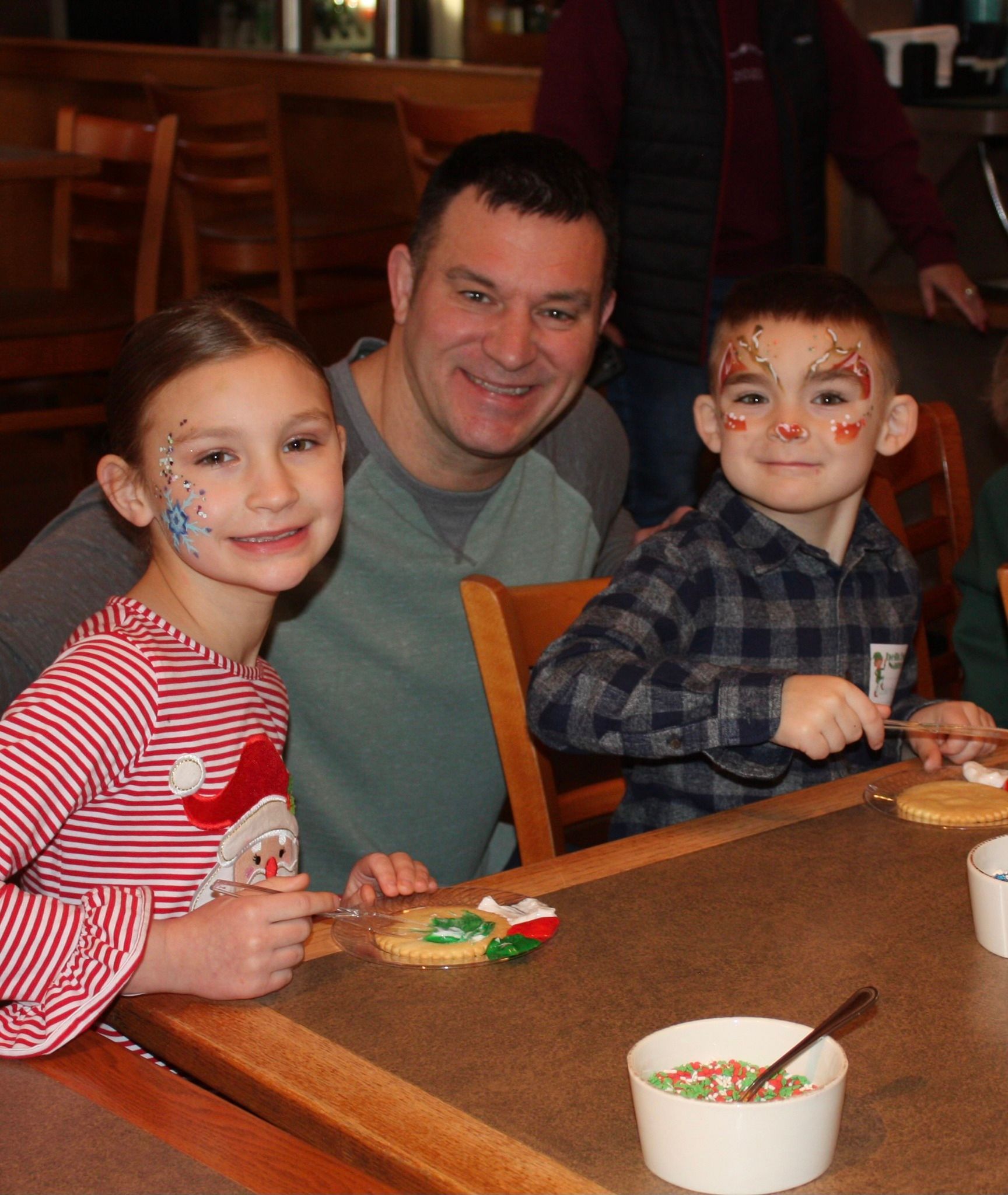 Man smiles with two children at a table decorating cookies. Face paint, sprinkles, holiday shirts.