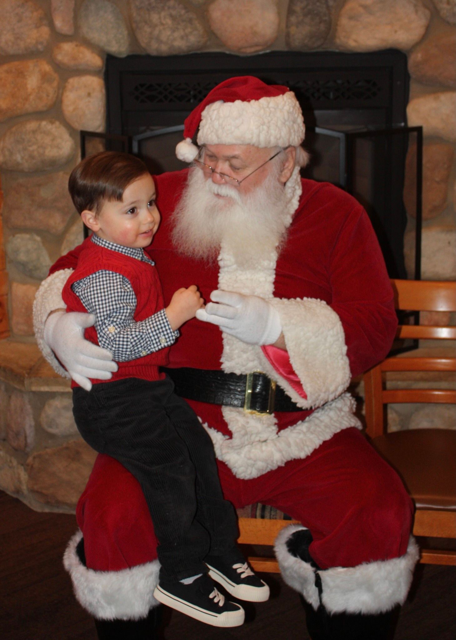 Child sitting on Santa's lap, smiling. Santa wearing a red suit with a white beard, near a fireplace.