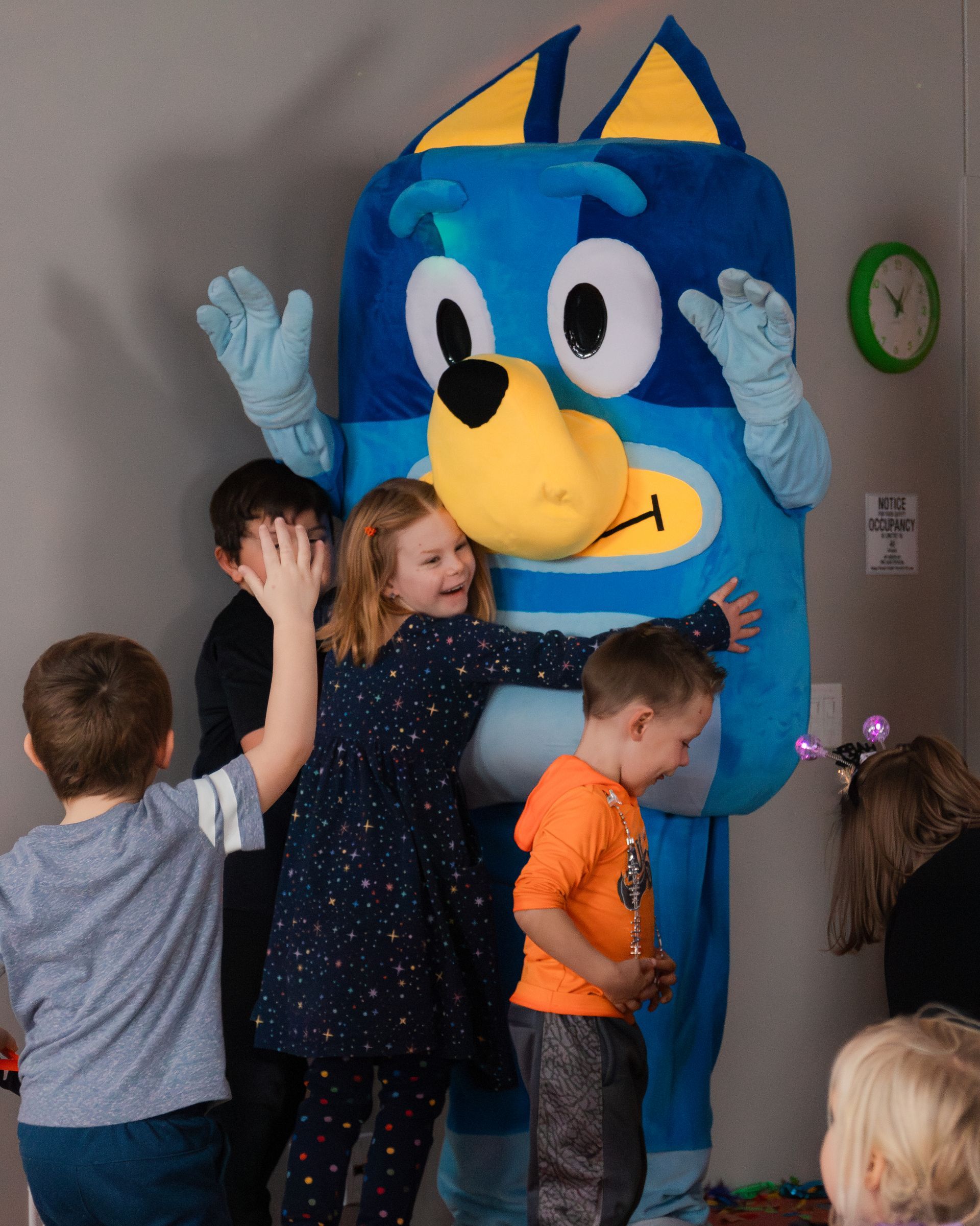 Children hugging Bluey mascot at a party. Blue and yellow colors.