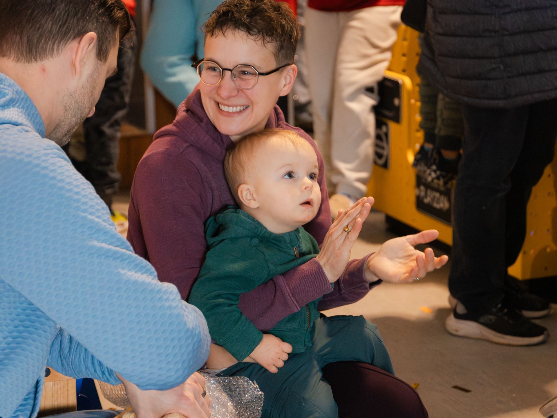 Woman smiles, holding a baby who gestures. A man is next to them, looking at the baby. Indoors setting.
