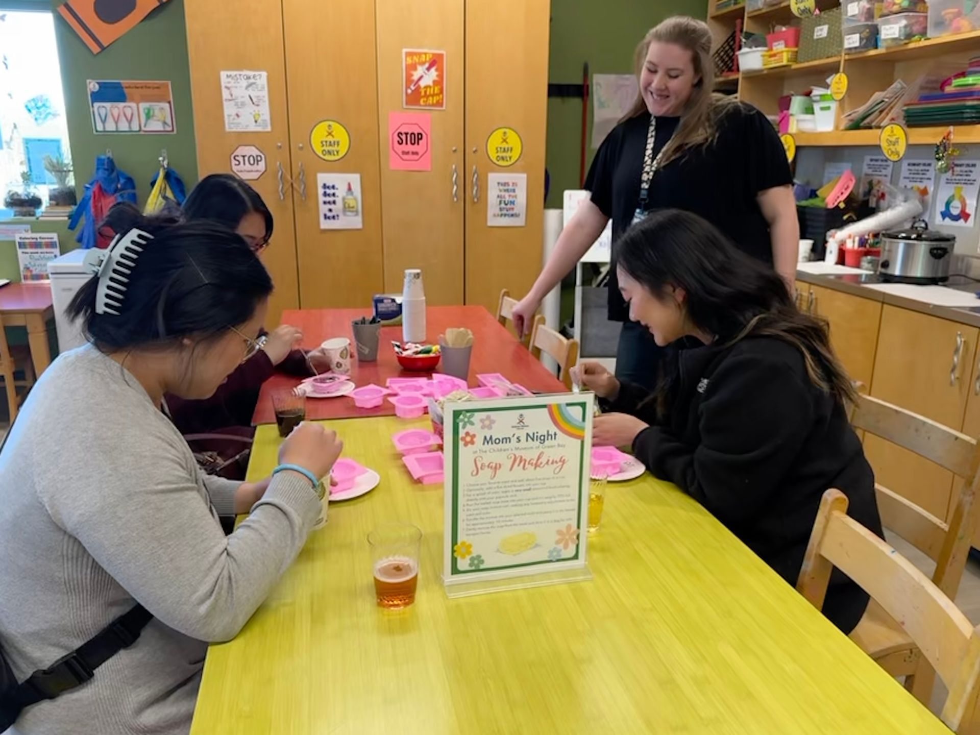 Four people sit at a bright yellow table in a classroom, working on a craft project with small pink silicone molds.