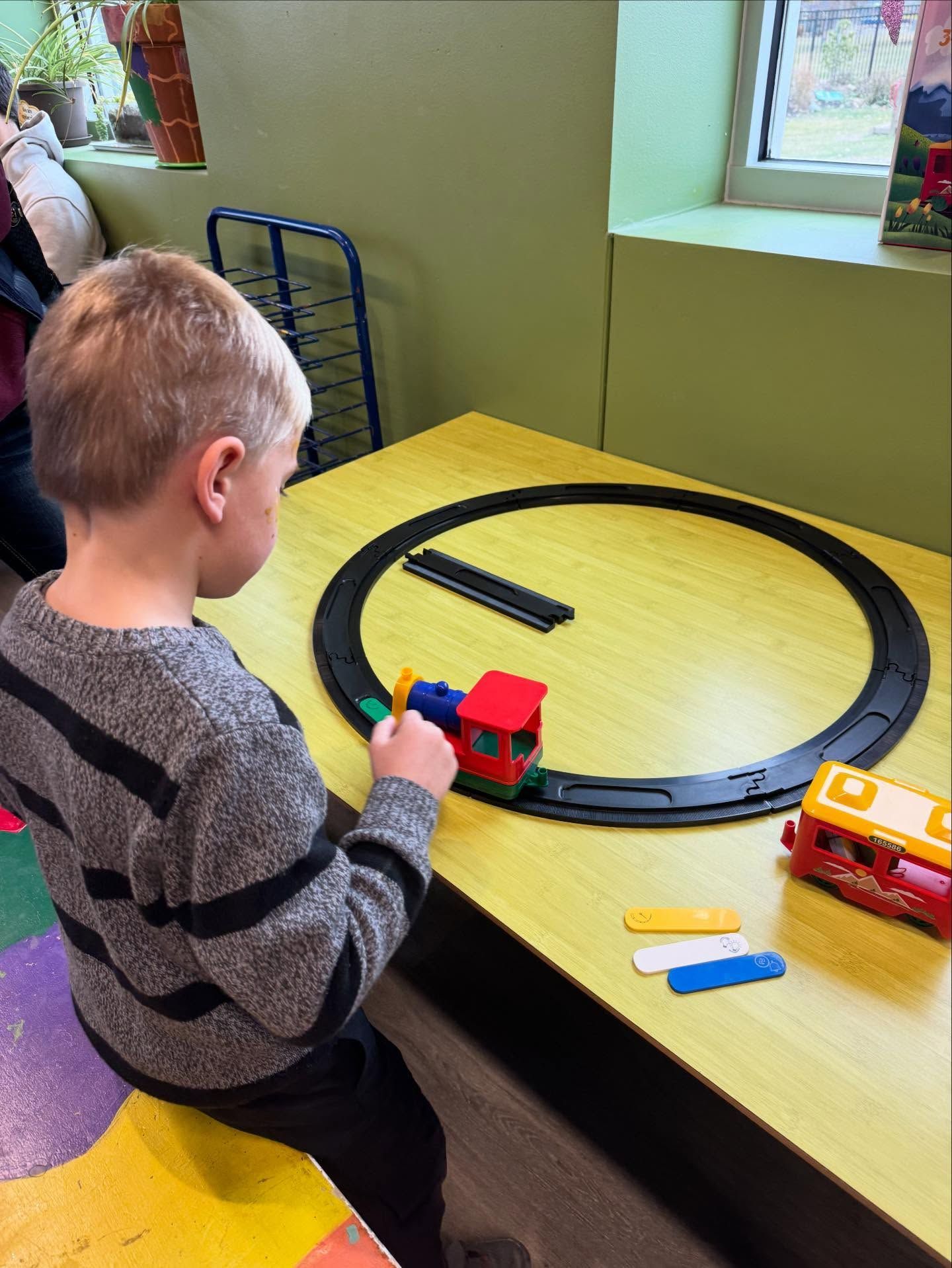 Boy playing with a toy train on a circular track. Yellow table, light green walls.