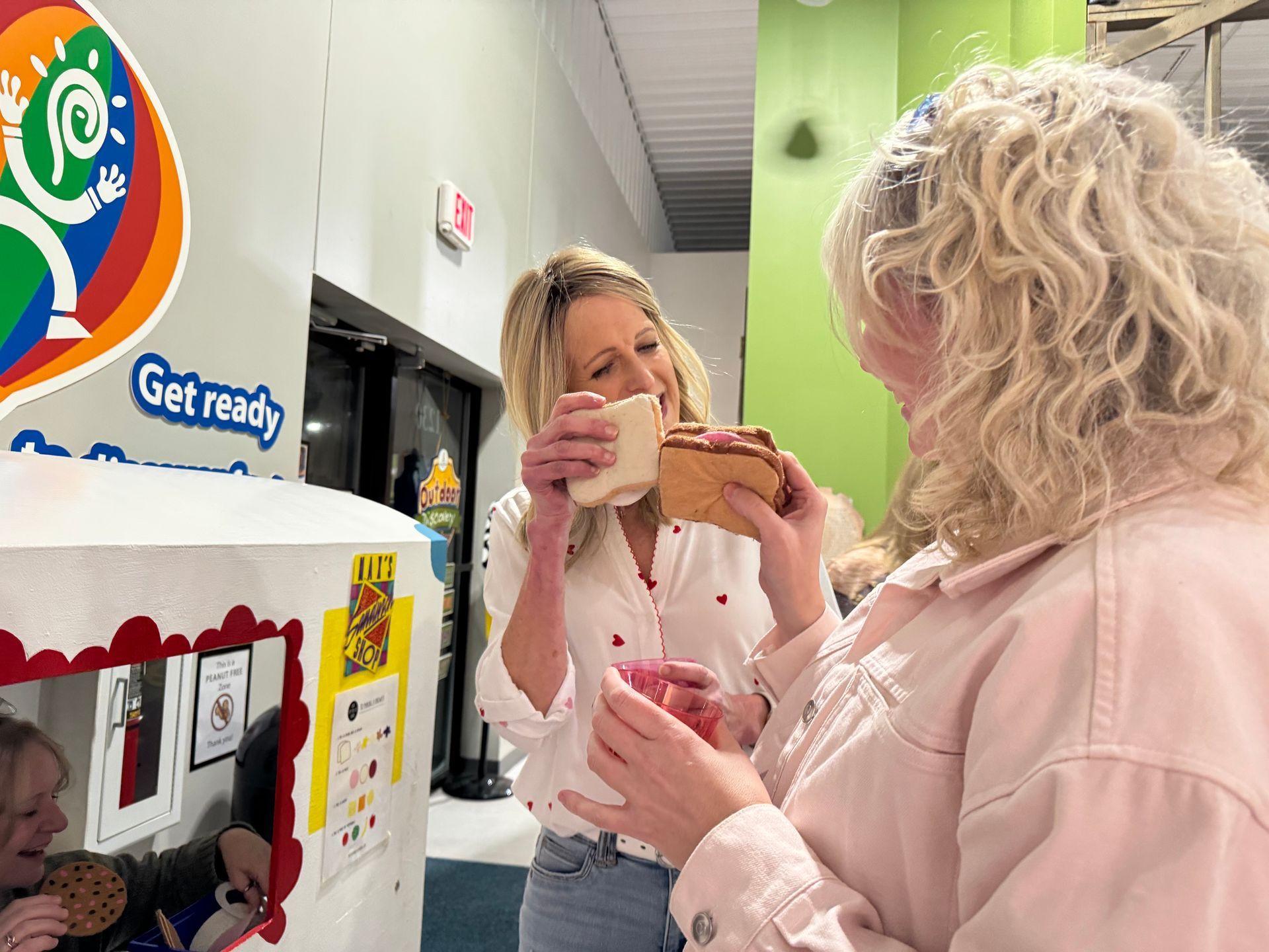 Two people stand indoors holding sandwiches; a child inside a small play structure eats a cookie in the background.