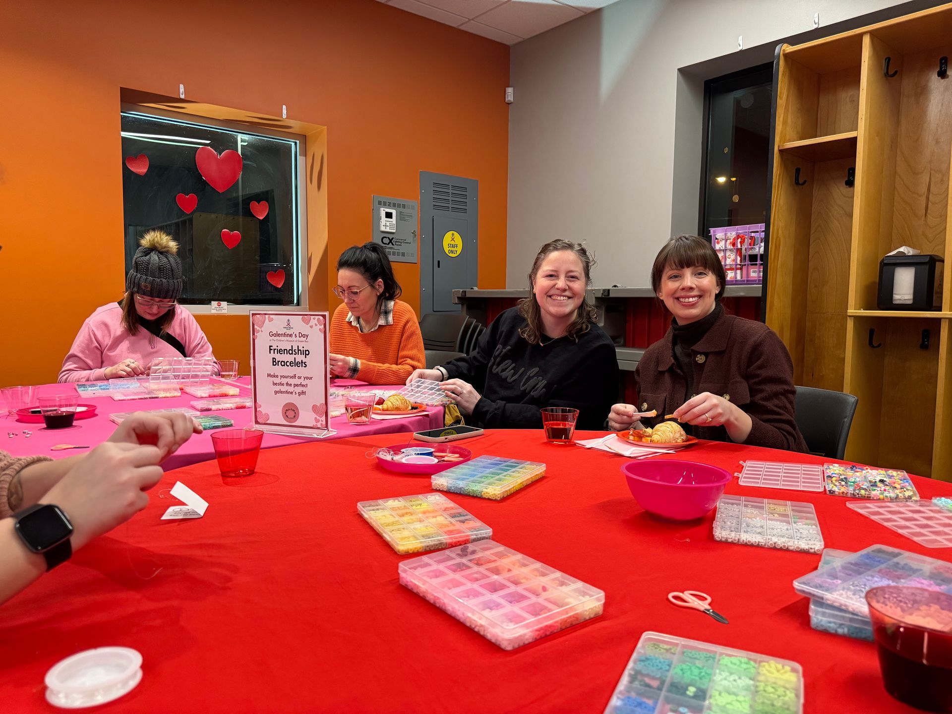 Four people sit around a large red table crafting with colorful beads in a bright, decorated room.