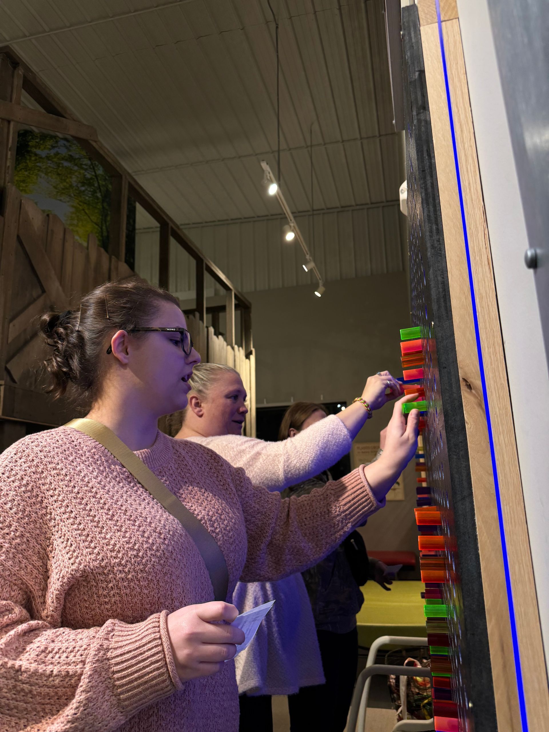 Two people in knit sweaters interact with a colorful, illuminated wall display in an indoor setting.