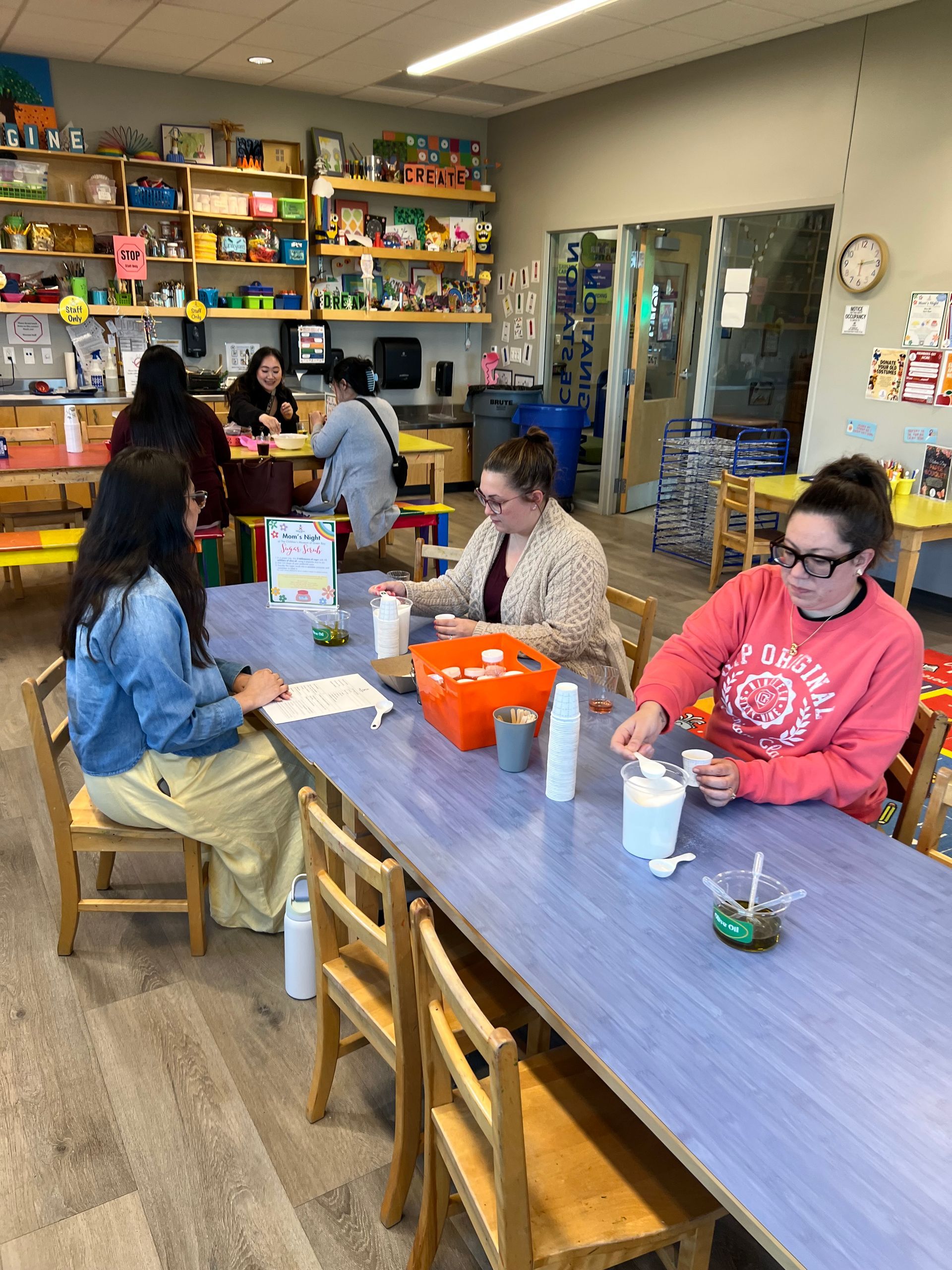 Four people sit at a blue table in a classroom, engaged in an activity with small white containers and supplies.