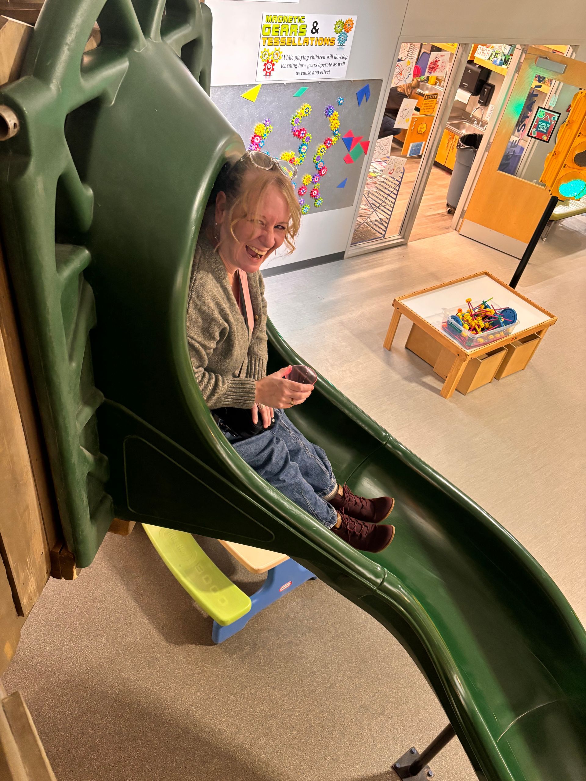 A person smiling while sitting at the top of a green indoor playground slide in a classroom.