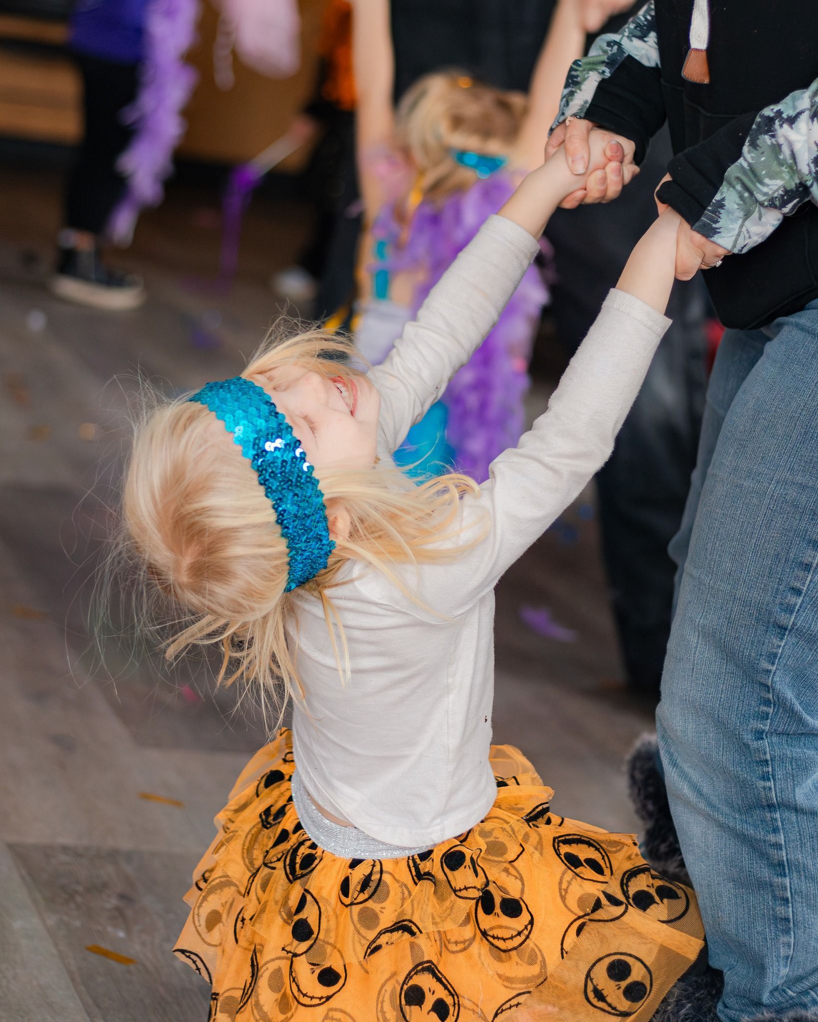 Girl dances, holding hands with adults, looking up with arms raised. She wears a blue headband, orange skirt with skulls.